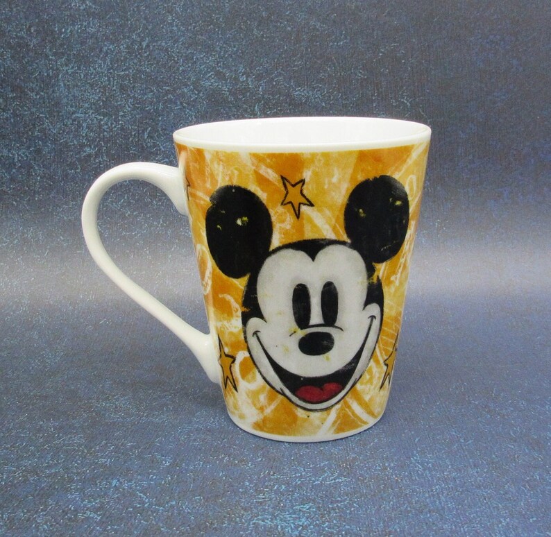 Puede incluir: Una taza de caf&eacute; de cer&aacute;mica amarilla con una imagen de dibujos animados de Mickey Mouse. La taza tiene un asa blanca y un contorno negro alrededor de la imagen.