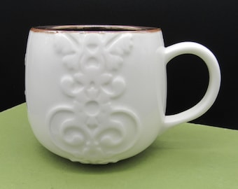 May include: White ceramic mug with a decorative embossed floral design. The mug has a rounded shape, a curved handle, and a gold-colored rim. The mug is sitting on a green surface against a black background.