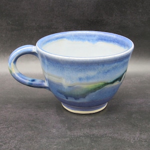 May include: A ceramic mug featuring a wide, rounded bowl shape with a single curved handle. The exterior displays a blended glaze of deep blue, light blue, and hints of green, while the interior is finished in a smooth, off-white glaze.