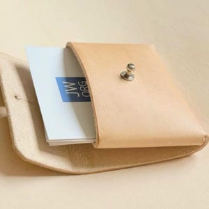 Könnte beinhalten: Ein hellbrauner Kartenhalter aus Leder mit einem silberfarbenen Metallknopfverschluss. Das Etui ist leicht geöffnet und gibt den Blick auf einen Stapel weißer Visitenkarten frei, von denen eine ein blaues Quadratlogo mit dem Text JW ORG trägt.