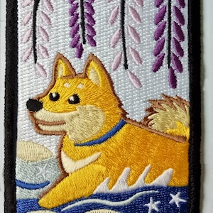 Puede incluir: Parche bordado con un perro Shiba Inu de pelaje amarillo, collar azul y cola esponjosa. El fondo incluye flores de glicina y elementos de agua estilizados. El parche tiene un borde negro.