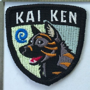 May include: Embroidered patch with the text "KAI KEN" in white block letters. The patch features a cartoon dog with black fur, orange markings, and a red tongue. The background is light green and blue.
