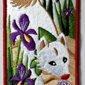 Op de afbeelding: Geborduurde patch met een wit dier met bruine oren en een rode neus, omringd door paarse irissen en groen gebladerte. Een bruine vogel en een paarse slak zijn ook aanwezig. De patch heeft een rode rand.