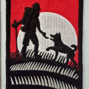 May include: Embroidered patch featuring a black silhouette of a person with a backpack and a dog on a grassy hill, set against a white moon and a red sky. The patch has a black border.