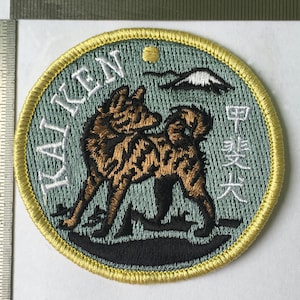 Könnte beinhalten: Gestickter Aufnäher mit einem braunen und schwarzen Hund mit dem Text "KAI KEN" und japanischen Schriftzeichen. Der Aufnäher hat einen hellgrünen Hintergrund mit einem gelben Rand und einer Bergillustration.