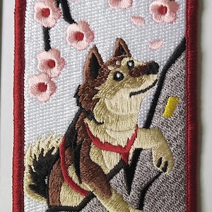 Può includere: Toppa ricamata raffigurante un cane marrone e beige con un'imbracatura rossa, che si arrampica su una superficie grigia. Fiori di ciliegio rosa adornano l'angolo in alto a sinistra. La toppa ha un bordo rosso.