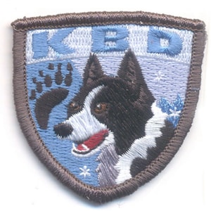 May include: Embroidered patch featuring a husky dog with black and white fur, a red tongue, and brown eyes. The patch has a blue background with the letters "KBD" and a bear paw print. The border is brown.
