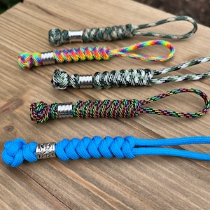 May include: Five different paracord keychains with metal beads. The keychains are in various colors and patterns, including camouflage, rainbow, black and neon green, and solid blue.