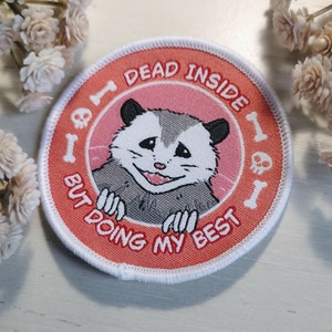 May include: A pink and white embroidered patch with a cartoon possum and the text "I'm dead inside but doing my best".