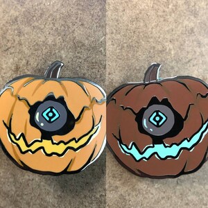 May include: Two enamel pins shaped like jack-o'-lanterns with a single eye in the center. One is orange with a yellow grin and the other is brown with a teal grin.
