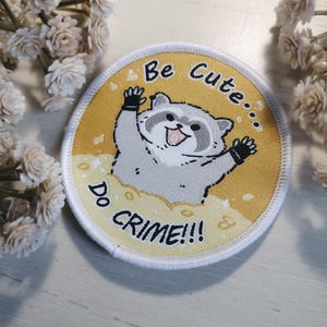Puede incluir: Un parche redondo amarillo y blanco con un mapache de dibujos animados que lleva una camisa negra y guantes negros. El mapache sonríe y tiene un fondo blanco. El texto en el parche dice "Be Cute... Do Crime!!"