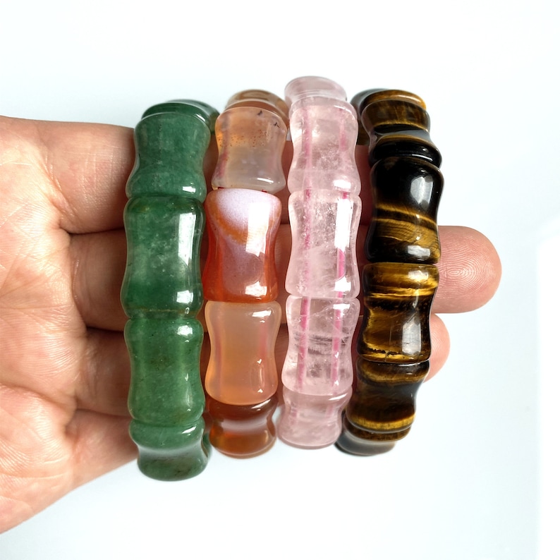 Op de afbeelding: Vier edelsteenarmbanden in groen, oranje, roze en bruin. Elke armband heeft een bamboe-achtig ontwerp, met afgeronde segmenten. De edelstenen worden in een hand tegen een witte achtergrond gehouden.