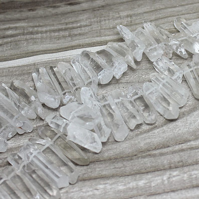 K&ouml;nnte beinhalten: Nahaufnahme von klaren Quarz-Kristallperlen. Die Kristalle sind grob geschnitten und unregelm&auml;&szlig;ig geformt, aufgef&auml;delt zu einer Kette oder einem Armband. Die durchscheinenden Kristalle werden auf einer Holzoberfl&auml;che pr&auml;sentiert.
