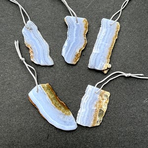 May include: Five blue lace agate pendants with a natural, rough edge and a silver cord for hanging. The pendants have a light blue colour with brown and gold accents. The pendants are irregularly shaped and vary in size.