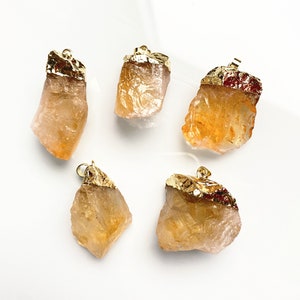 Pode incluir: Cinco pingentes de cristal de quartzo bruto de cor laranja com topos banhados a ouro. Os cristais são de forma irregular e têm uma textura natural e áspera.