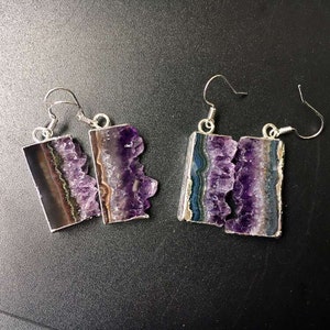 Puede incluir: Un par de pendientes de plata con detalles de cristal de amatista. Los cristales son de color morado intenso y tienen una textura natural y rugosa. Los pendientes son un accesorio &uacute;nico y elegante.