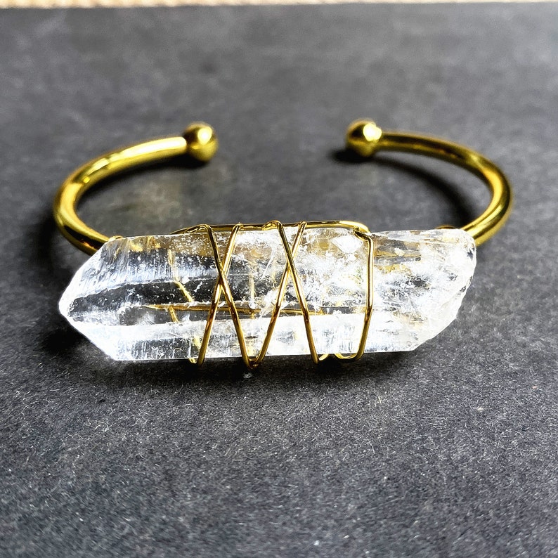 May include: A gold wire wrapped clear quartz crystal is attached to a gold bangle bracelet.