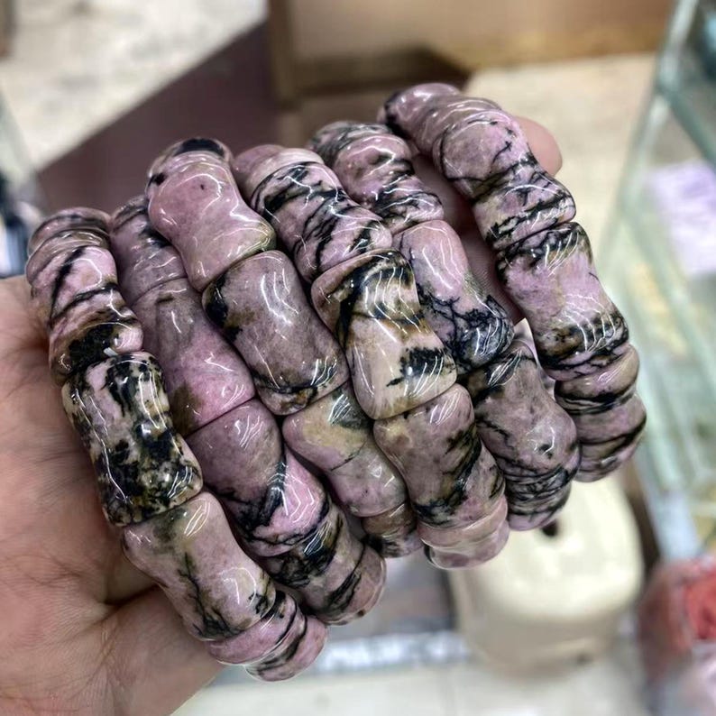 Op de afbeelding: Verschillende roze en zwarte stenen armbanden worden getoond. De armbanden zijn gemaakt van gepolijste, afgeronde stenen met een gemarmerd uiterlijk. De stenen zijn voornamelijk roze met zwarte adering. De armbanden zijn op elkaar gestapeld, wat hun ontwerp en kleurvariaties benadrukt.