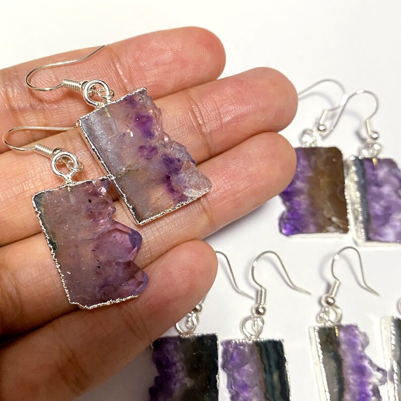 Puede incluir: Un par de pendientes de plata con detalles de piedra de amatista p&uacute;rpura. Las piedras tienen una forma irregular y un aspecto natural y bruto. Los pendientes son una pieza llamativa y ser&iacute;an una gran adici&oacute;n a cualquier colecci&oacute;n de joyas.