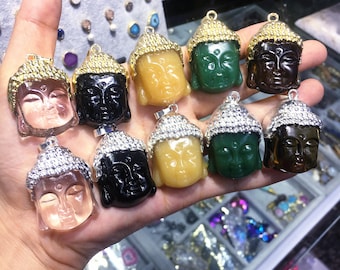 May include: A collection of ten Buddha head pendants, each carved from a different semi-precious stone. The pendants are displayed on a hand, with the stones ranging in color from clear to black, green, and brown.