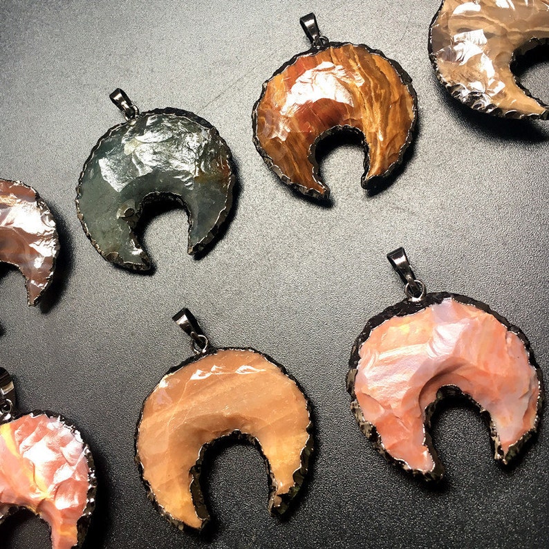 May include: A collection of ten crescent moon shaped pendants made from agate. The pendants are all different colors and patterns, including brown, pink, green, and white. Each pendant has a silver bail for hanging.