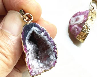 May include: Two gemstone pendants with gold-colored metal accents. One pendant is a purple and white geode slice with a gold-colored edge. The other pendant is a pink geode slice with a gold-colored edge.