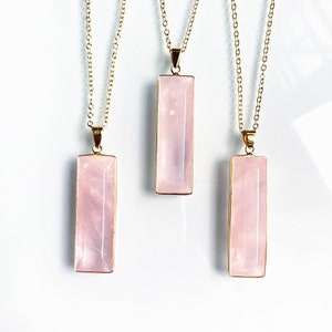 May include: Three gold necklaces with pink rectangular crystal pendants. The necklaces are on a white background.