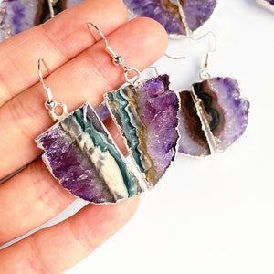 Peut inclure: Une paire de boucles d'oreilles en cristal d'améthyste avec des crochets argentés. Les boucles d'oreilles sont faites d'améthyste brute avec une forme naturelle irrégulière. L'améthyste est d'une couleur violet foncé avec des inclusions blanches et vertes.