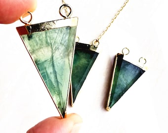 May include: Three gold-rimmed green fluorite triangle pendants on a gold chain. The pendants are different sizes and have a natural, cloudy appearance.