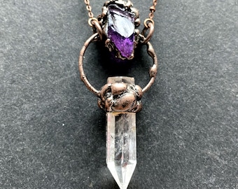 Puede incluir: Un collar de cobre con una punta de cristal de cuarzo transparente y un colgante de amatista púrpura. El colgante está sujeto en un círculo de alambre de cobre.