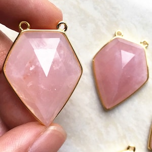 May include: Two pink rose quartz gemstone pendants with gold-toned metal settings. The pendants are shaped like a diamond and have a faceted design.