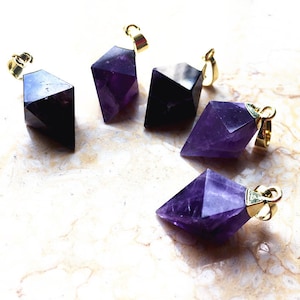 May include: Five faceted amethyst pendants with gold-colored bails. The amethyst crystals are a deep purple color and have a geometric shape. The pendants are displayed on a light-colored surface.