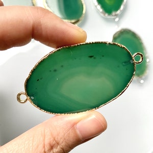 Puede incluir: Rebanada de ágata verde ovalada con un borde dorado y dos anillos de salto dorados. La ágata tiene un color verde claro con vetas verde oscuro.