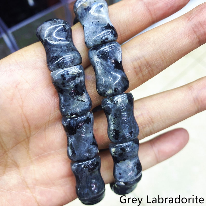 Op de afbeelding: Close-up van grijze labradoriet kralen. De kralen zijn cilindrisch met een licht zandlopervorm. De steen heeft een mix van grijze en zwarte tinten. De tekst "Grey Labradorite" is rechtsonder zichtbaar.