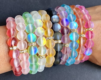 Könnte beinhalten: Ein Set mit 12 Armbändern aus schimmernden Glasperlen. Jedes Armband hat einen goldenen Abstandshalter mit einem klaren Strassstein.