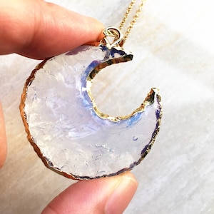 May include: A gold chain necklace with a crescent moon pendant made of clear iridescent glass. The pendant has a gold rim.
