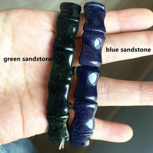 Op de afbeelding: Twee zandstenen armbanden, &eacute;&eacute;n groen en &eacute;&eacute;n blauw, elk bestaande uit cilindrische kralen. De groene armband heeft een gespikkelde uitstraling, terwijl de blauwe armband een glad, reflecterend oppervlak heeft. Tekst op de afbeelding luidt "green sandstone" en "blue sandstone".