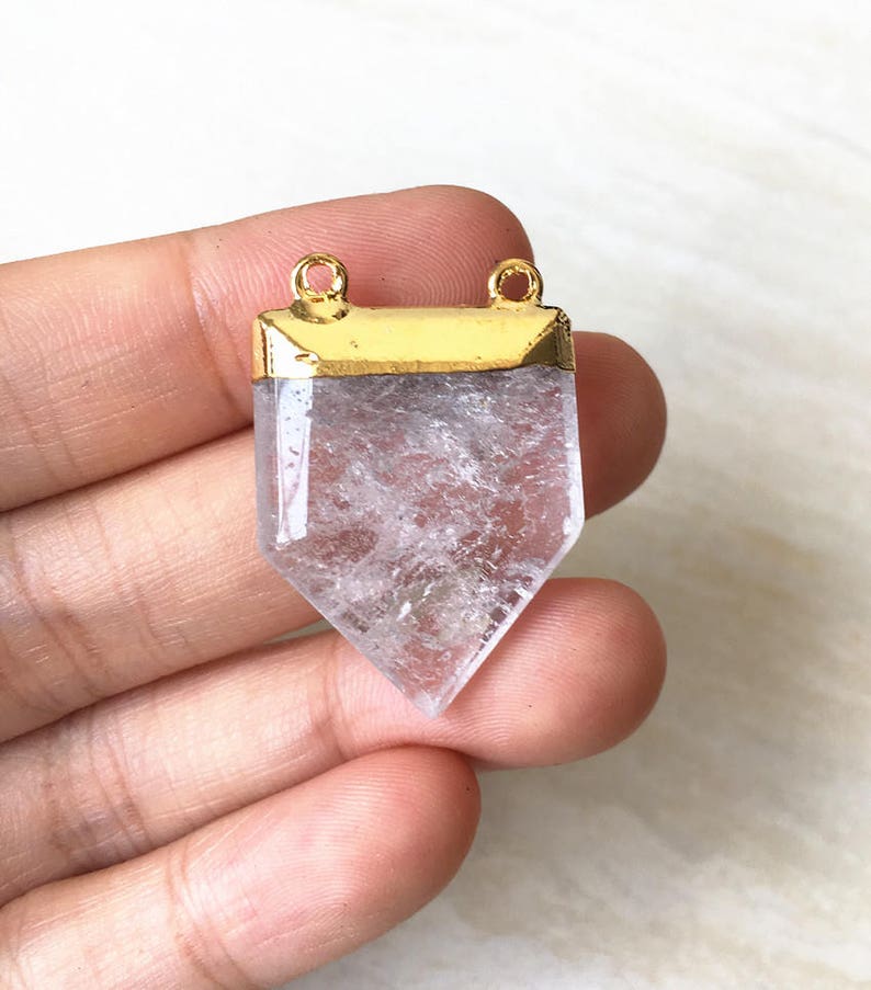 Puede incluir: Un colgante de cristal de cuarzo transparente con una parte superior chapada en oro y dos anillos de salto. El cristal est&aacute; facetado y tiene una punta puntiaguda.