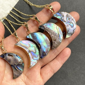 Pode incluir: Cinco colares de ouro com pingentes de lua crescente de concha de abalone iridescente. Os colares estão sobrepostos em uma mão.