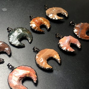 May include: A collection of eight crescent moon shaped pendants made from natural agate. Each pendant has a unique pattern of brown, orange, and gray colors. The pendants are accented with a black metal trim.