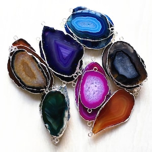 Op de afbeelding: Assortiment van agaatplakken met zilverkleurige metalen accenten. De plakken zijn in verschillende kleuren, waaronder blauw, paars, groen, roze, bruin en zwart.