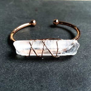 May include: A rose gold wire-wrapped clear quartz crystal bangle bracelet. The crystal is wrapped in rose gold wire and is set on a rose gold bangle.