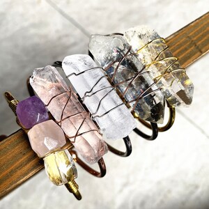 May include: Five crystal bracelets with copper wire wrapped around them. The crystals are clear, pink, and purple. The bracelets are stacked on top of each other.