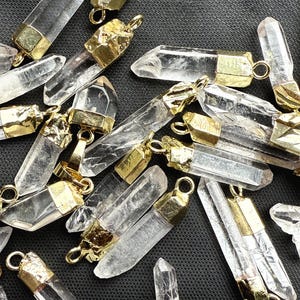 May include: A collection of clear quartz crystal pendants with gold-toned metal caps. The crystals are faceted and have a pointed tip. The pendants are perfect for jewelry making or crafting.