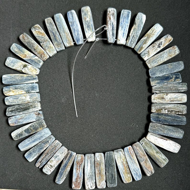 Pode incluir: Um colar de contas retangulares em tons de azul, cinza e marrom. As contas s&atilde;o enfiadas com um fio fino e transparente, formando uma forma circular. O fundo &eacute; preto liso.