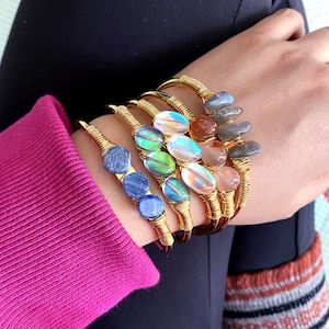 May include: Five gold wire wrapped bracelets with different colored gemstone beads. The bracelets feature iridescent, blue, green, and pink stones.