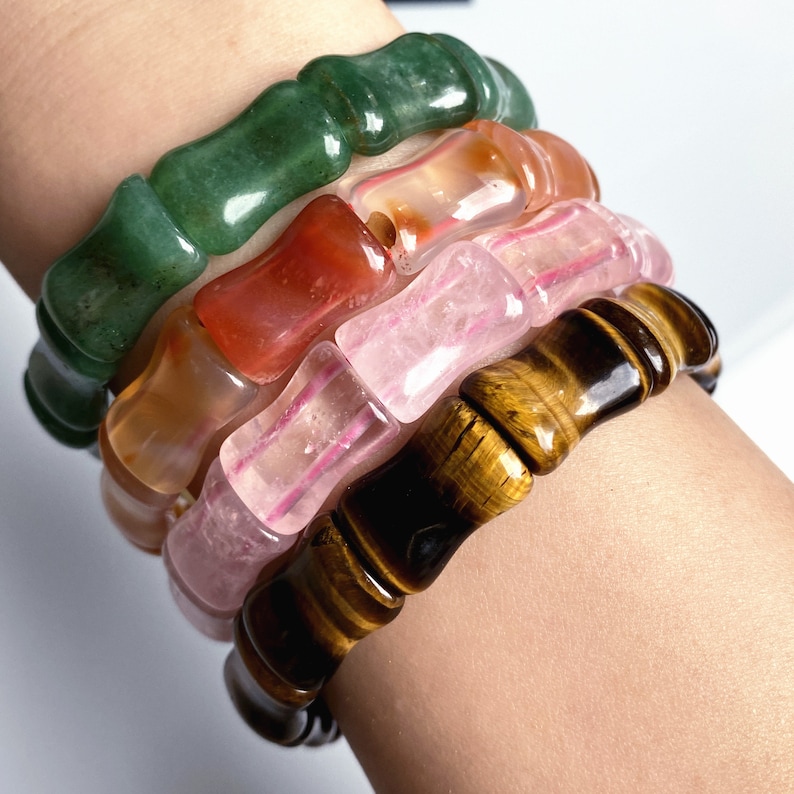 Op de afbeelding: Vier edelsteenarmbanden in groen, oranje, roze en bruin zijn op een pols gestapeld. Elke armband heeft een unieke kleur en patroon, met een gepolijste, gladde afwerking. De armbanden zijn gemaakt van jade, agaat, rozenkwarts en tijgeroog.