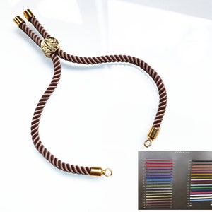 Può includere: Un braccialetto in corda intrecciata marrone con chiusura regolabile dorata e un disco decorativo dorato. Il braccialetto è mostrato su uno sfondo bianco. Una cartella colori con 25 colori diversi di corda intrecciata è mostrata sotto il braccialetto.