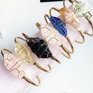 May include: A set of six gold wire wrapped bracelets featuring different natural crystal stones. The stones include clear quartz, blue kyanite, orange calcite, green aventurine, black obsidian, and pink rose quartz.