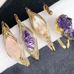Op de afbeelding: Vier gouden draadgewikkelde armbanden met verschillende gekleurde edelstenen. De armbanden hebben een roze, een paarse, een lichtroze en een paarse amethist steen.